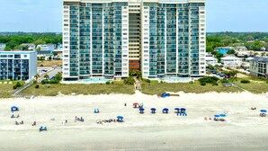 A high-rise hotel with multiple balconies, a pool area, and a beach with umbrellas.