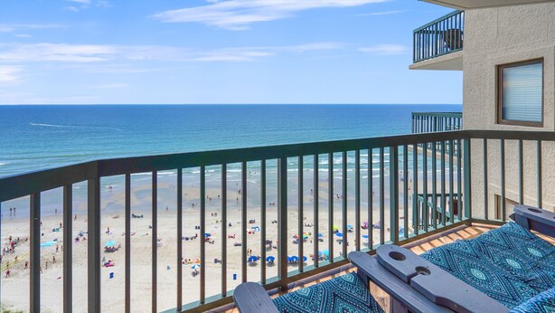 A balcony with a view of a beach, wooden lounge chairs, and a patterned cushion.