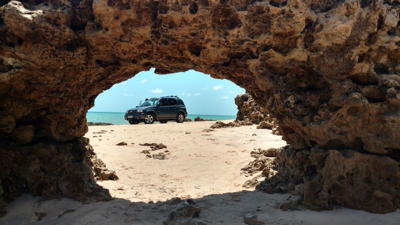 Um veículo estacionado por um arco rochoso numa praia arenosa, com vista clara para o oceano.