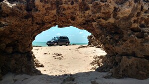 Un véhicule est garé sous une arche rocheuse, sur une plage de sable, d’où l’on a une vue dégagée sur l’océan.