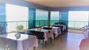 Un restaurant amb taules preparades per sopar, grans finestres que donen vista al mar i una zona de balcó amb caires.