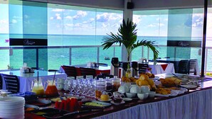 Taula de buffet amb diferents plats i begudes, davant d'un gran finestral amb vista al mar.