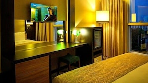 Habitación de hotel con una cama, un escritorio con televisor, una lámpara y una ventana con cortinas.