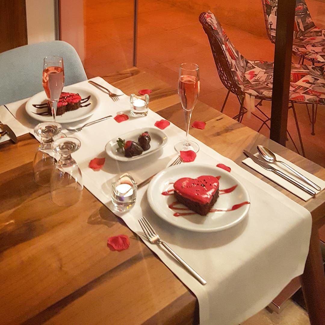 A romantic dinner setting with a table set for two, featuring a heart-shaped dessert and rose petals.
