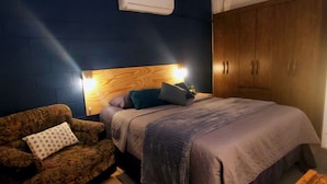 A bedroom with a bed, a wooden headboard, a nightstand, a lamp, a wardrobe, and a patterned armchair. - Alojamiento El Salvador: Villa Madero - Hotel Boutique (San Salvador)
