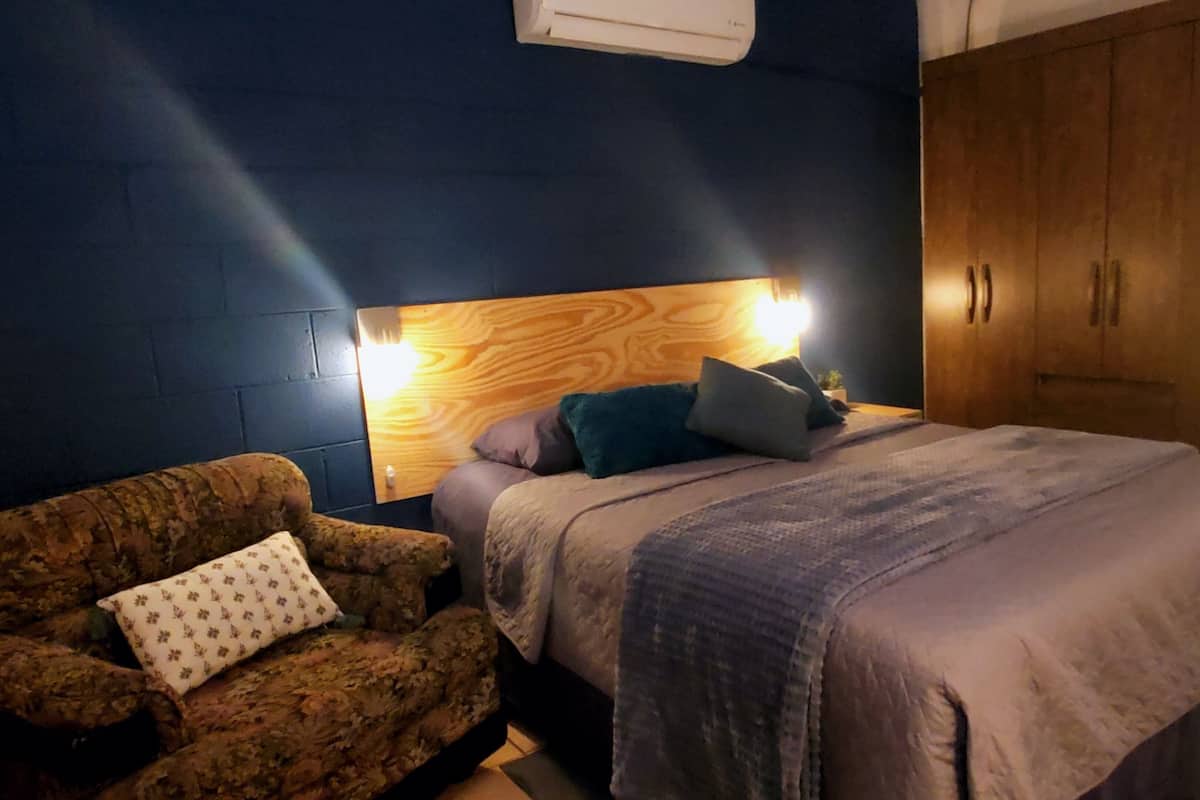 A bedroom with a bed, a wooden headboard, a nightstand, a lamp, a wardrobe, and a patterned armchair.