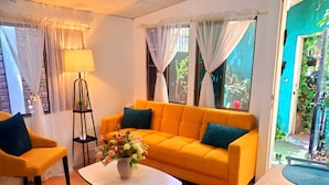 A room with a yellow sofa, a small round table, and a lamp. There are two chairs, a rug, and a window with curtains. - Alojamiento El Salvador: Villa Madero - Hotel Boutique (San Salvador)