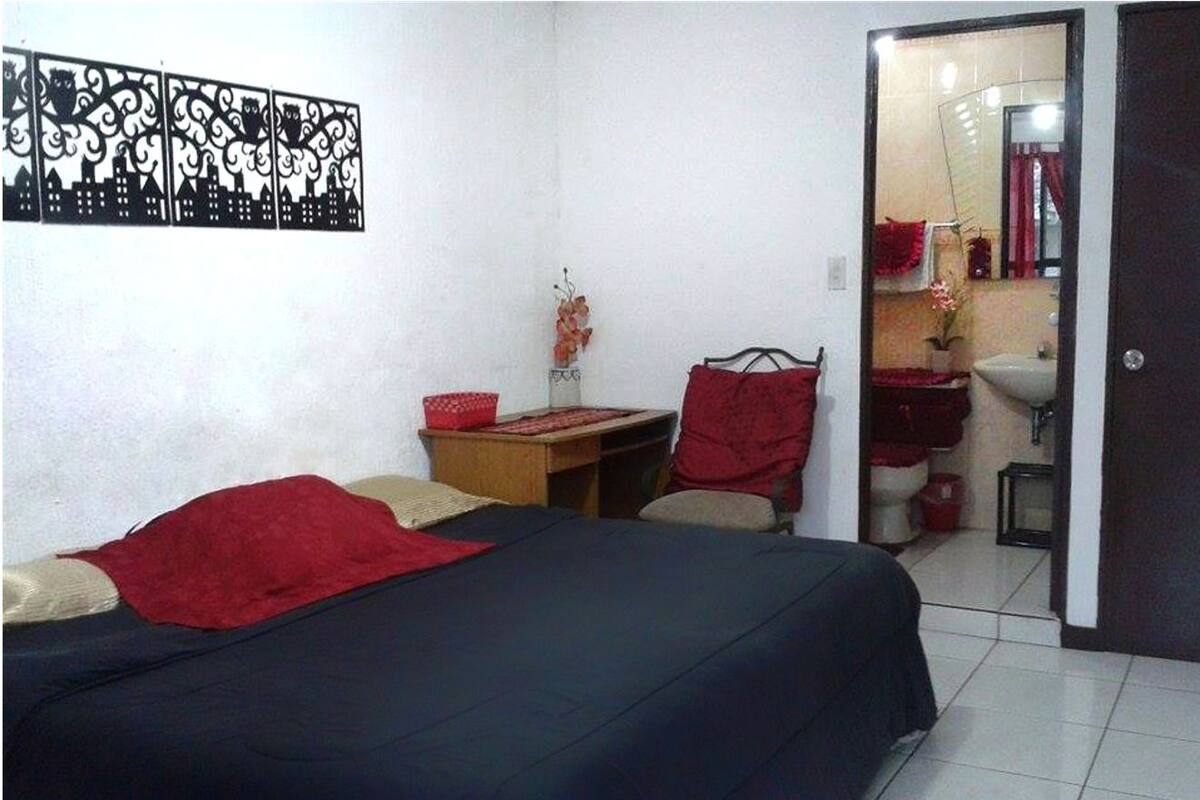A bedroom with a bed, a desk, a chair, and a bathroom visible through an open door.