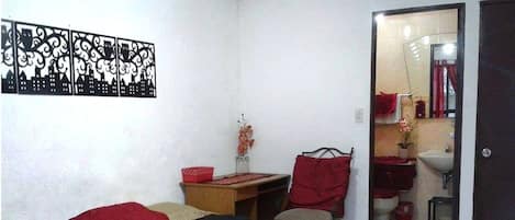 Une chambre à coucher avec un lit, un bureau, une chaise et une salle de bain visible à travers une porte ouverte.