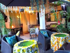 An outdoor seating area with wicker furniture, covered by a canopy, and decorated with hanging plants and lights.