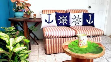 Un espacio de sala acogedor con un sofá a rayas, almohadas con motivos náuticos, una mesita con un tapete verde y plantas en macetas.