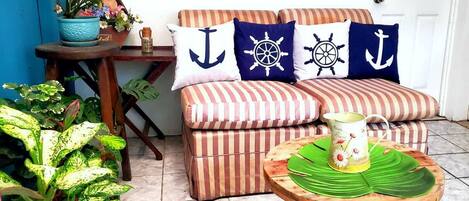 Un espacio de sala acogedor con un sofá a rayas, almohadas con motivos náuticos, una mesita con un tapete verde y plantas en macetas.