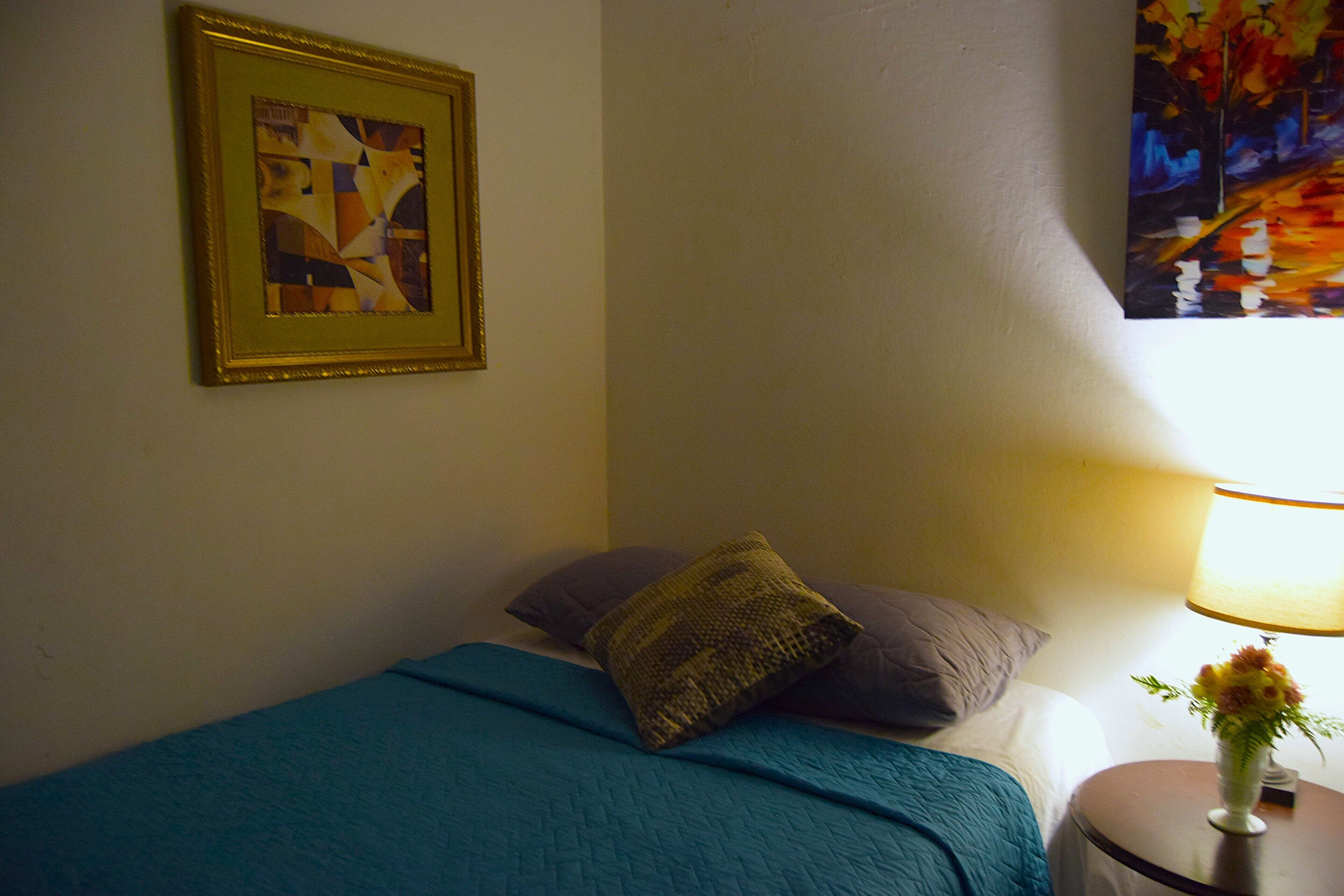 A bedroom with a bed, pillows, a framed abstract painting, a lamp, and a vase with flowers.