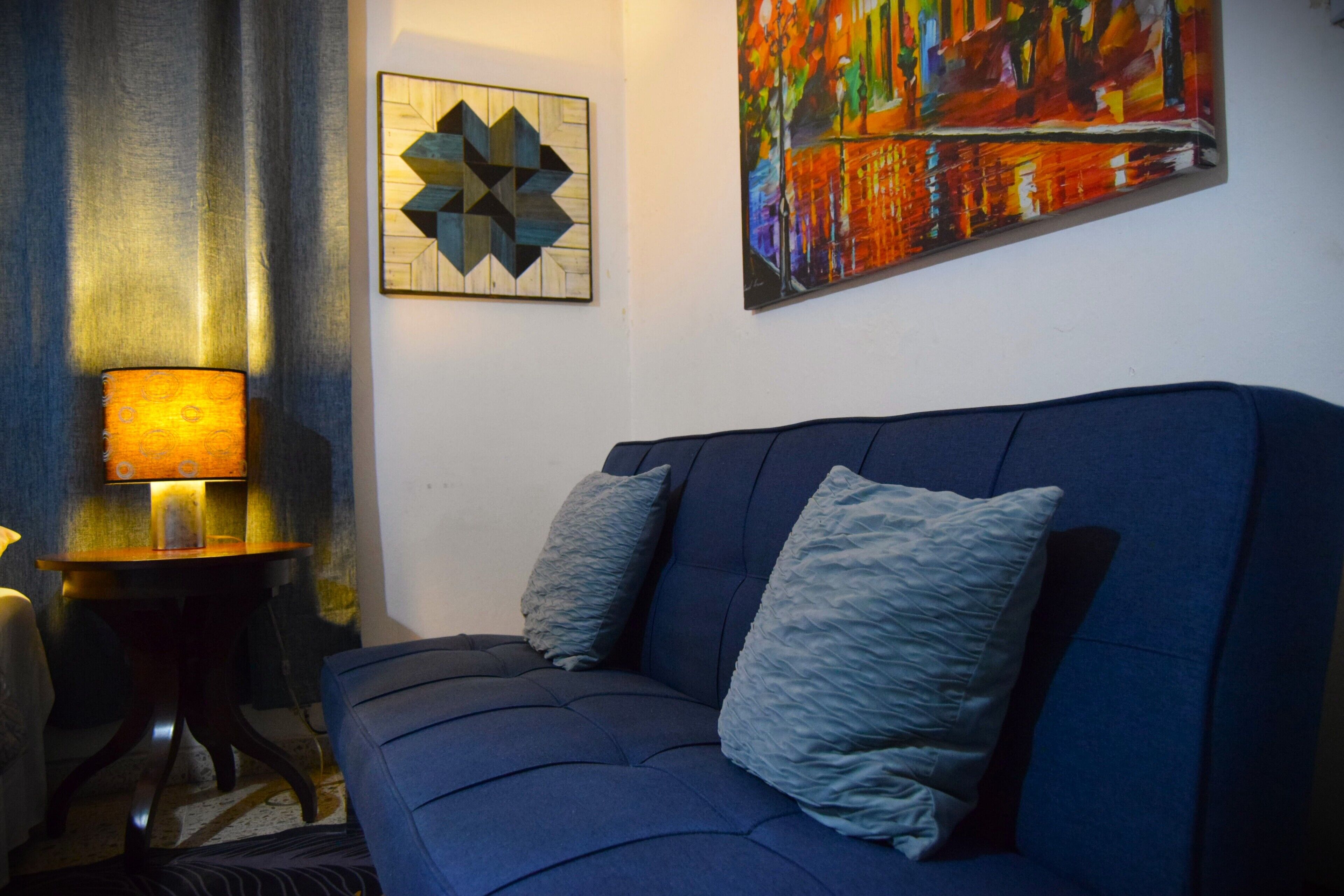 A modern living room with a blue sofa, two pillows, a lamp, and two artworks on the wall.
