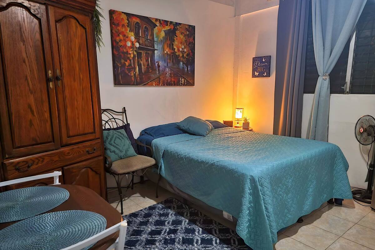 A bedroom with a bed, a chair, a table, a cabinet, a painting, a lamp, and a fan.