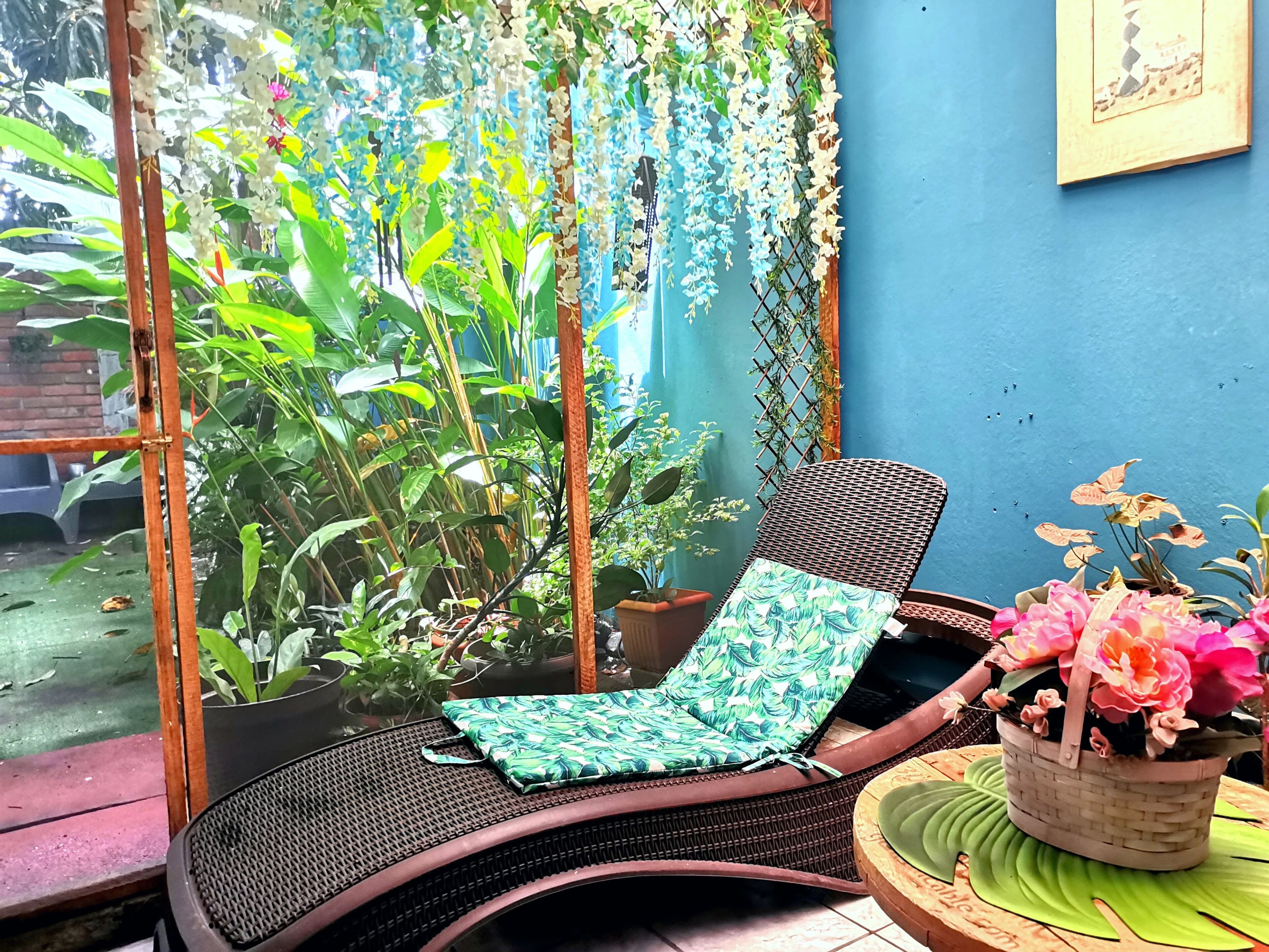 Un fauteuil de jardin en osier, doté d’un coussin à motifs de feuilles vertes, un pot de fleurs et une fenêtre donnant sur un paysage verdoyant.