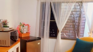 A compact kitchen with a microwave, refrigerator, and a wooden cabinet. There is a chair with a blue cushion and a window with sheer curtains. - Alojamiento El Salvador: Villa Madero - Hotel Boutique (San Salvador)