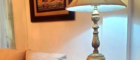 Una habitación con un sillón blanco, una mesa redonda negra con una lámpara, y una obra de arte enmarcada en la pared.