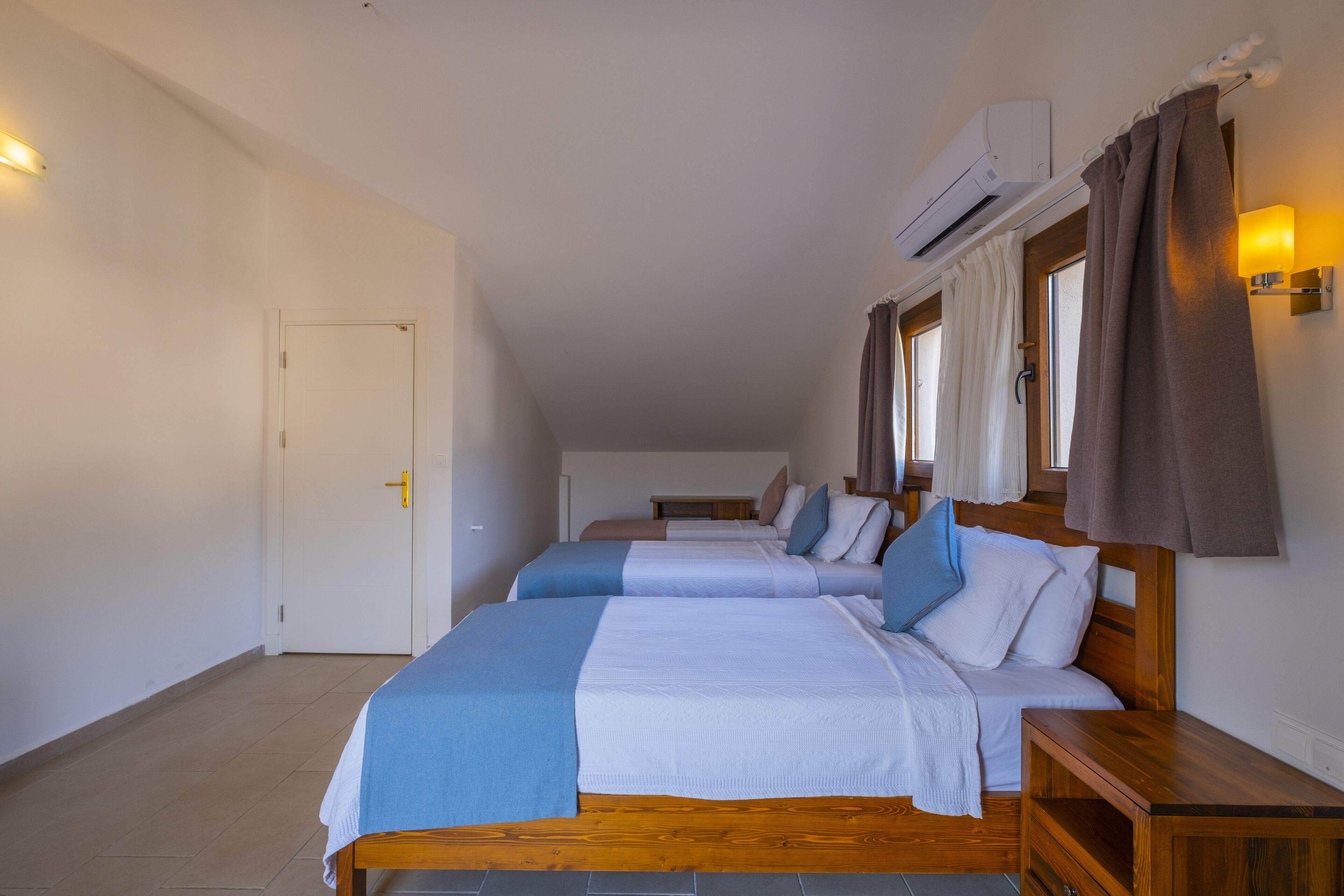 A hotel room with two beds, a wooden headboard, a nightstand, a wall-mounted air conditioner, and a window with curtains.