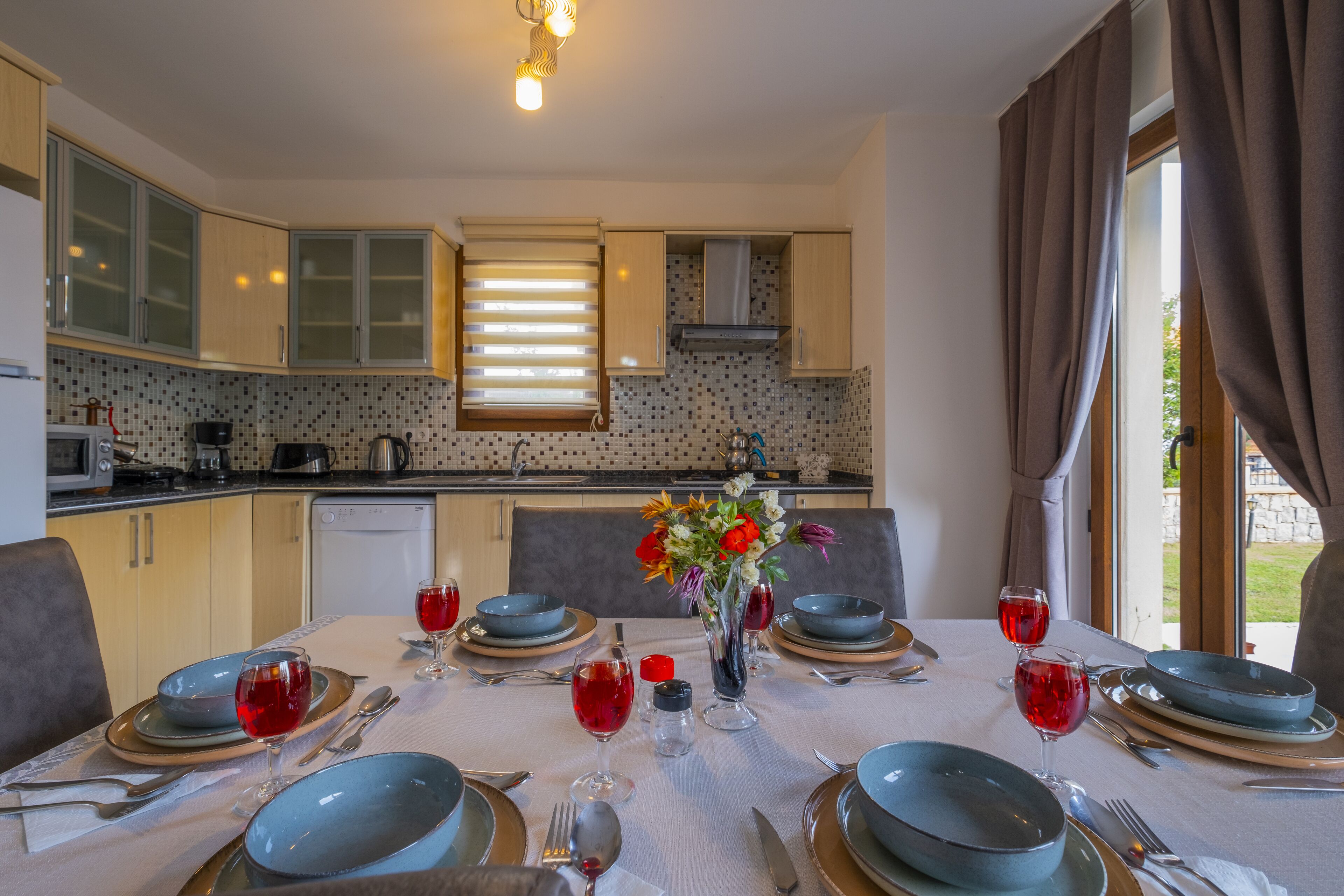 A dining area with a table set for six, featuring blue plates, silverware, and red wine glasses. A vase with flowers is placed on the table.