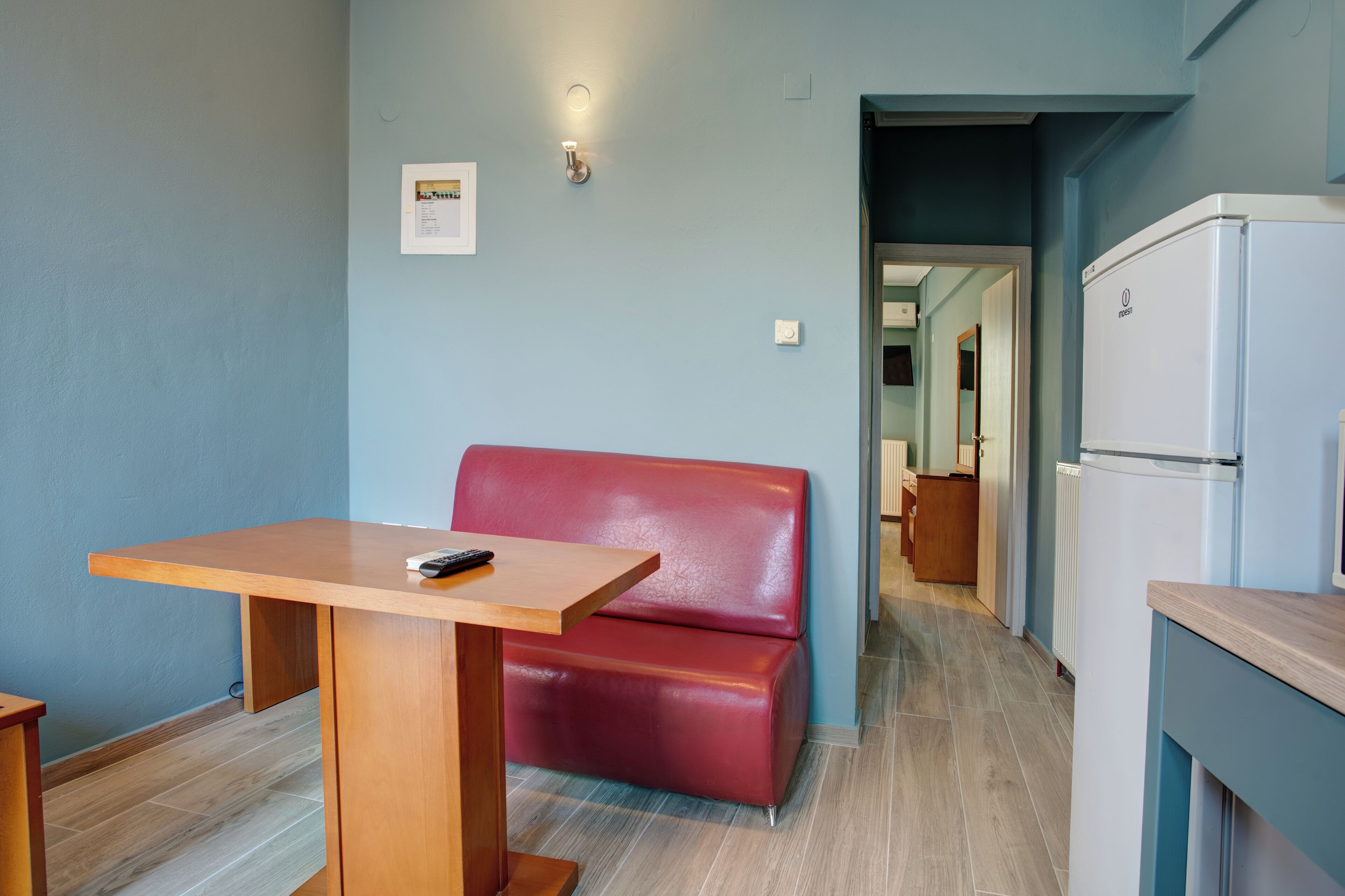 Una habitación pequeña con una mesa de madera, un banco rojo, un refrigerador blanco y un accesorio de iluminación en la pared.