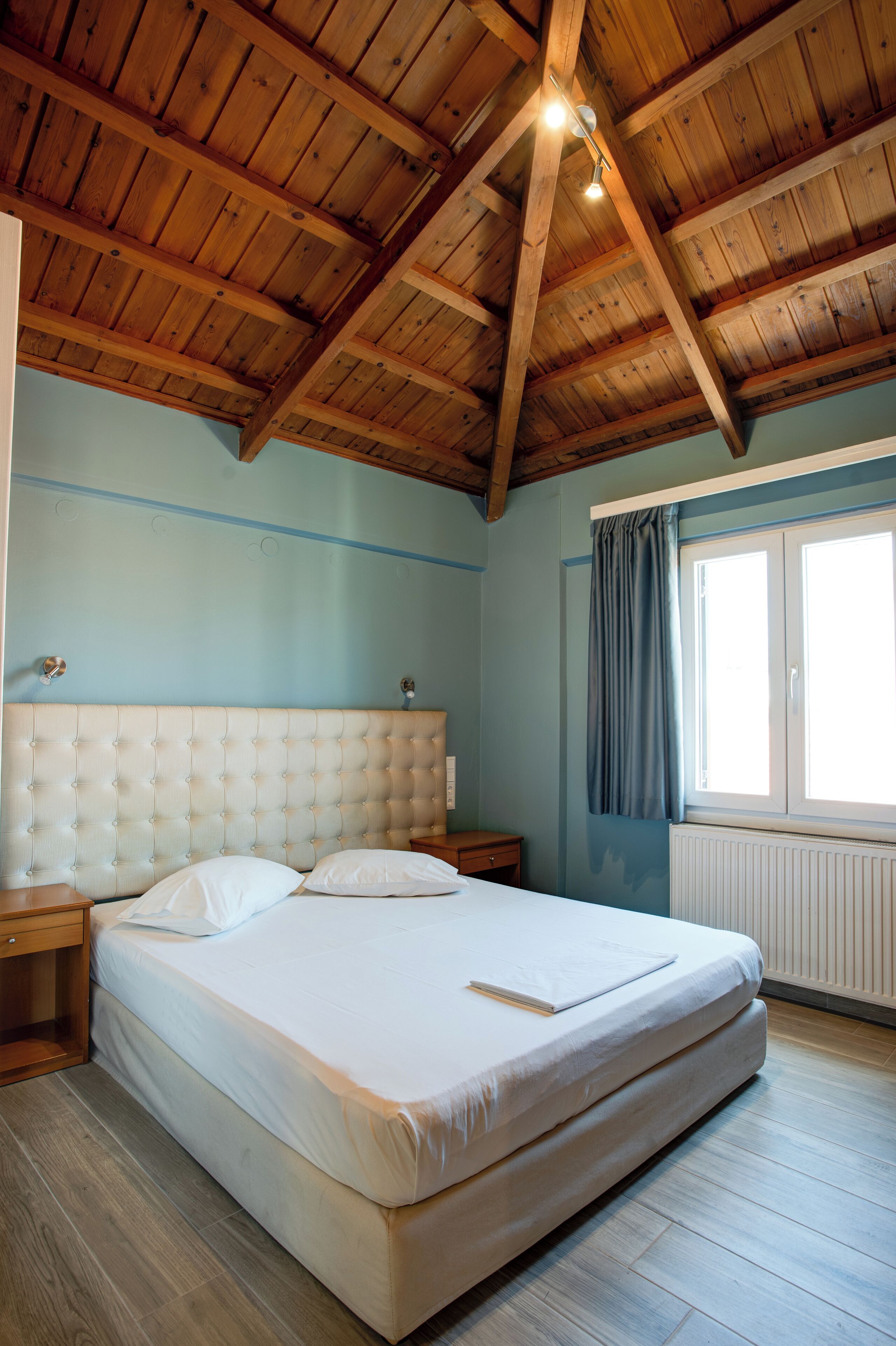 A bedroom with a wooden ceiling, a bed, a nightstand, a window with curtains, and a radiator.