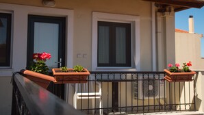 A balcony with two terracotta planters with flowers, a white chair, and a black metal railing. - Heliotrope The Studios (Lesvos)