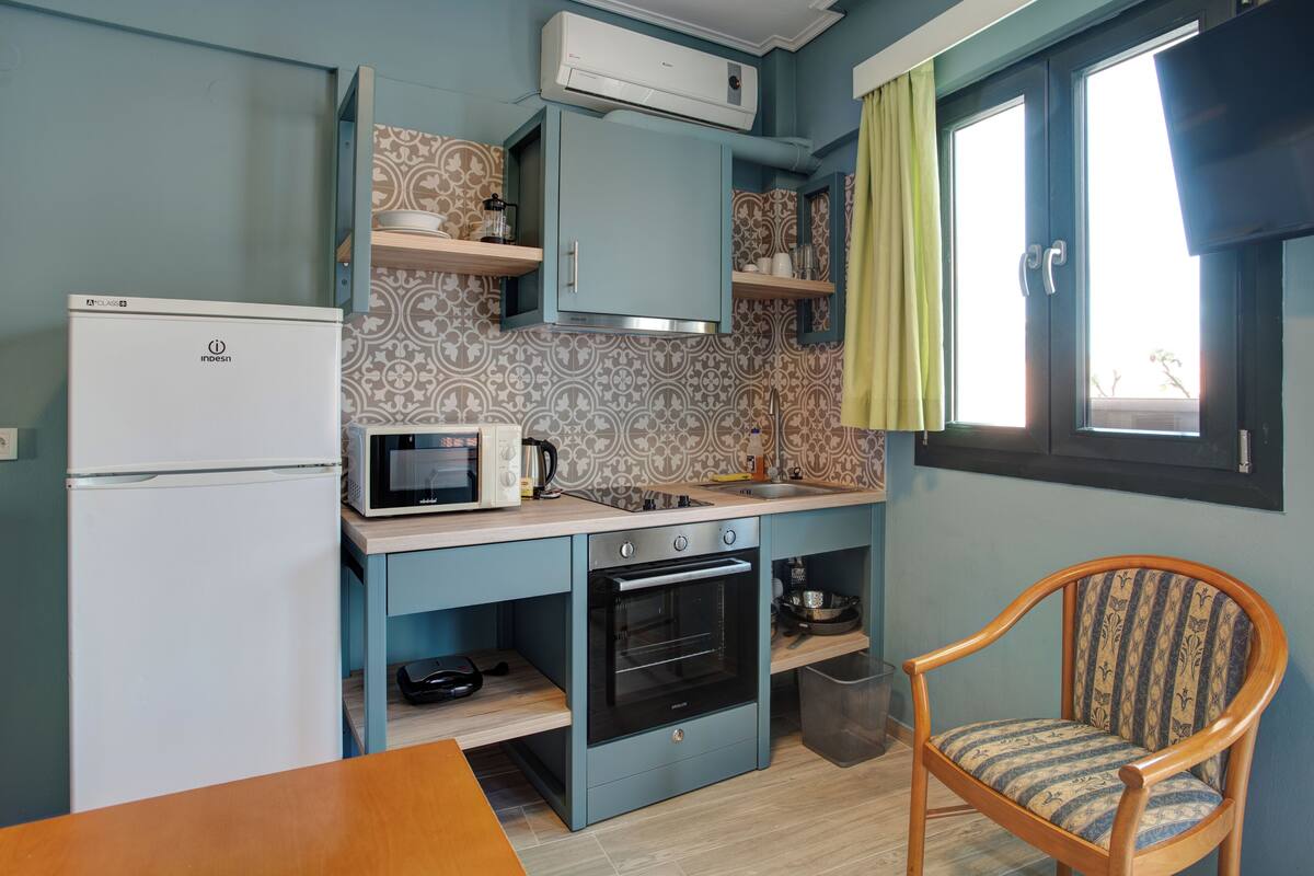 A compact kitchen with a refrigerator, microwave, oven, and a chair.