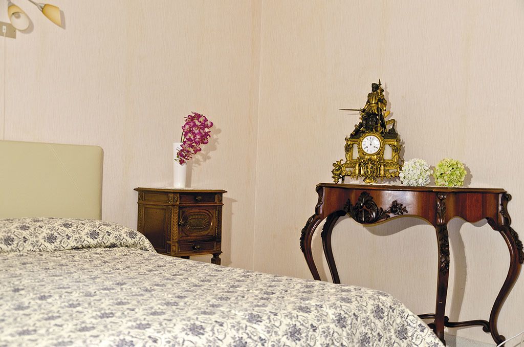 Ein Schlafzimmer mit einem Bett, einem Nachttisch mit Blumenvase sowie einem Tisch mit einer Uhr und Dekoration.