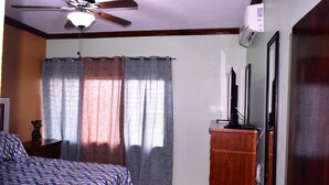 Un dormitorio con cama, ventilador de techo, cómoda, televisor y ventana con cortinas.