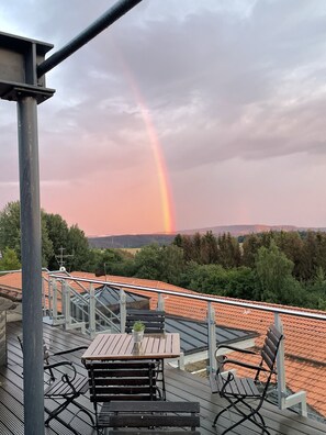 Een balkon met uitzicht op een regenboog, een pannendak en omliggend groen.