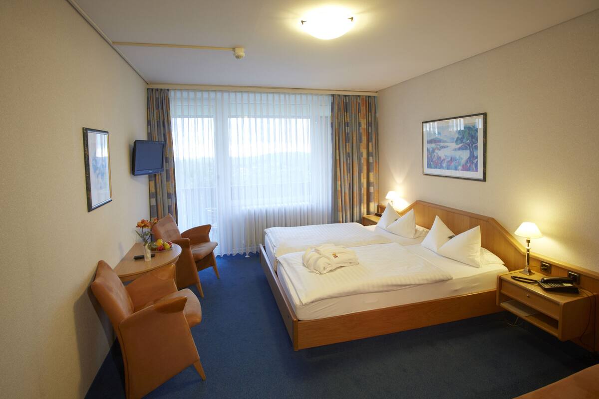 Een hotelkamer met een bed, nachtkastje, lamp, televisie en een bureau met stoelen.