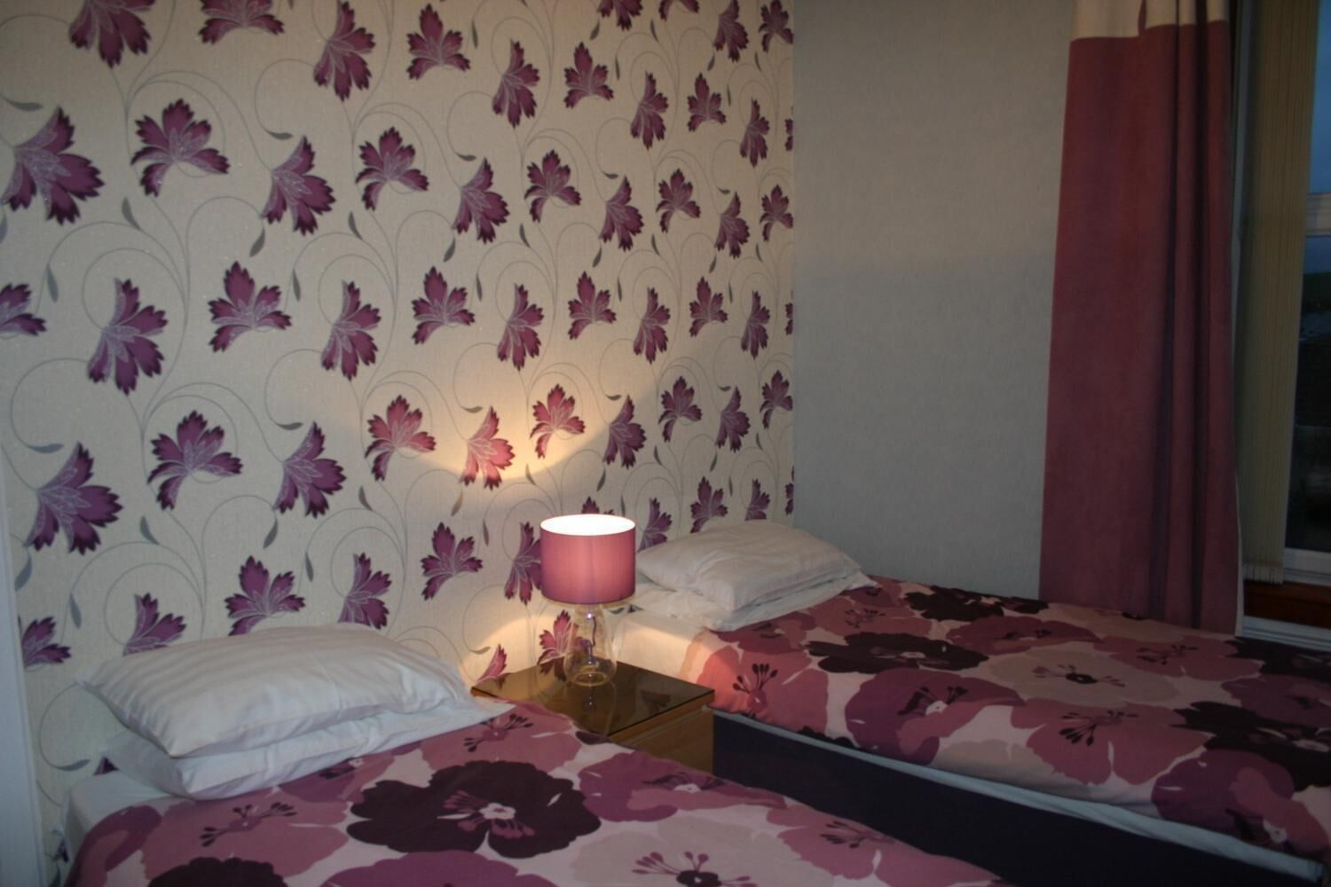 A bedroom with two beds, a nightstand, a lamp, and a window with curtains.