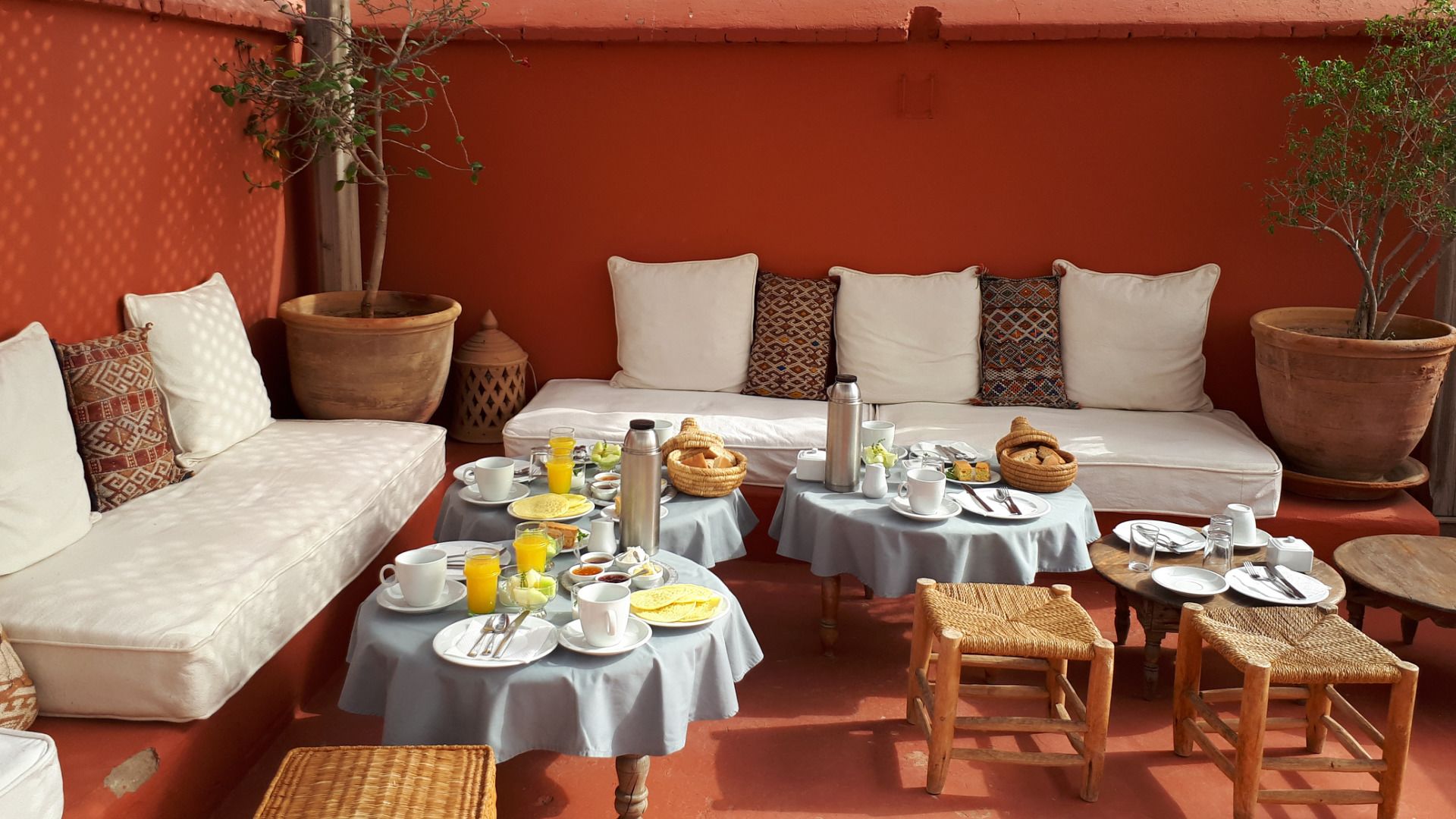 A cozy outdoor seating area with a sofa, two tables set for breakfast, and a small table with a basket of bread.
