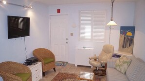 Uma sala de estar com cadeiras de vime, um sofá, uma mesinha, uma televisão e uma porta.