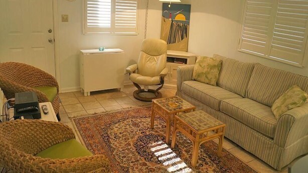 Una sala de estar con sofá, sillón y una mesita. Se ve una ventana con persianas, una lámpara y un cuadro en la pared.