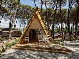 Una tienda de madera triangular con una mesa y dos sillas en su interior, rodeada de árboles.