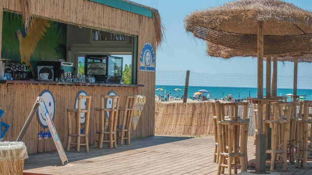 Un bar en la playa con fachada de madera, taburetes y un paraguas de paja.