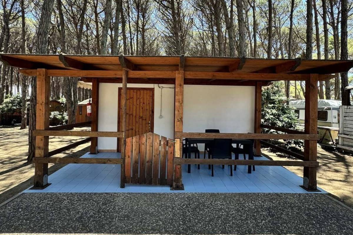 A wooden pergola structure with a table and chairs underneath, surrounded by a forested area.