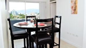 A dining area with a round table, black chairs, and a view of buildings and mountains through a large window. - New Kingston Luxury Apt The Bromptons (Kingston)