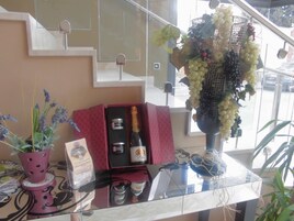 Zona de recepción de un hotel con una mesa de vidrio que muestra una botella de champán, un florero con uvas y una planta en maceta.