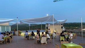 Un espace extérieur aménagé pour les repas, avec des tables et des chaises, une scène où se produit un artiste, et une vue panoramique en arrière-plan.
