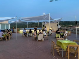 Un espacio de comedor al aire libre con mesas y sillas, un escenario con un artista y una vista panorámica de fondo.