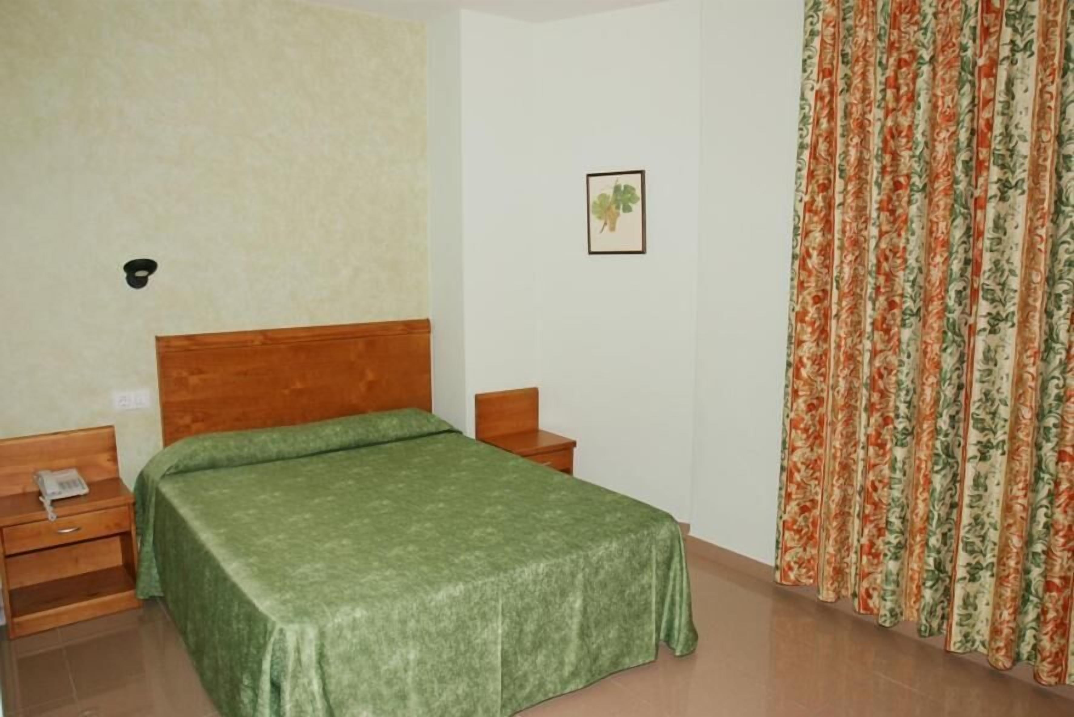 Quarto com cama, mesa de cabeceira, luminária na parede e cortina florida.