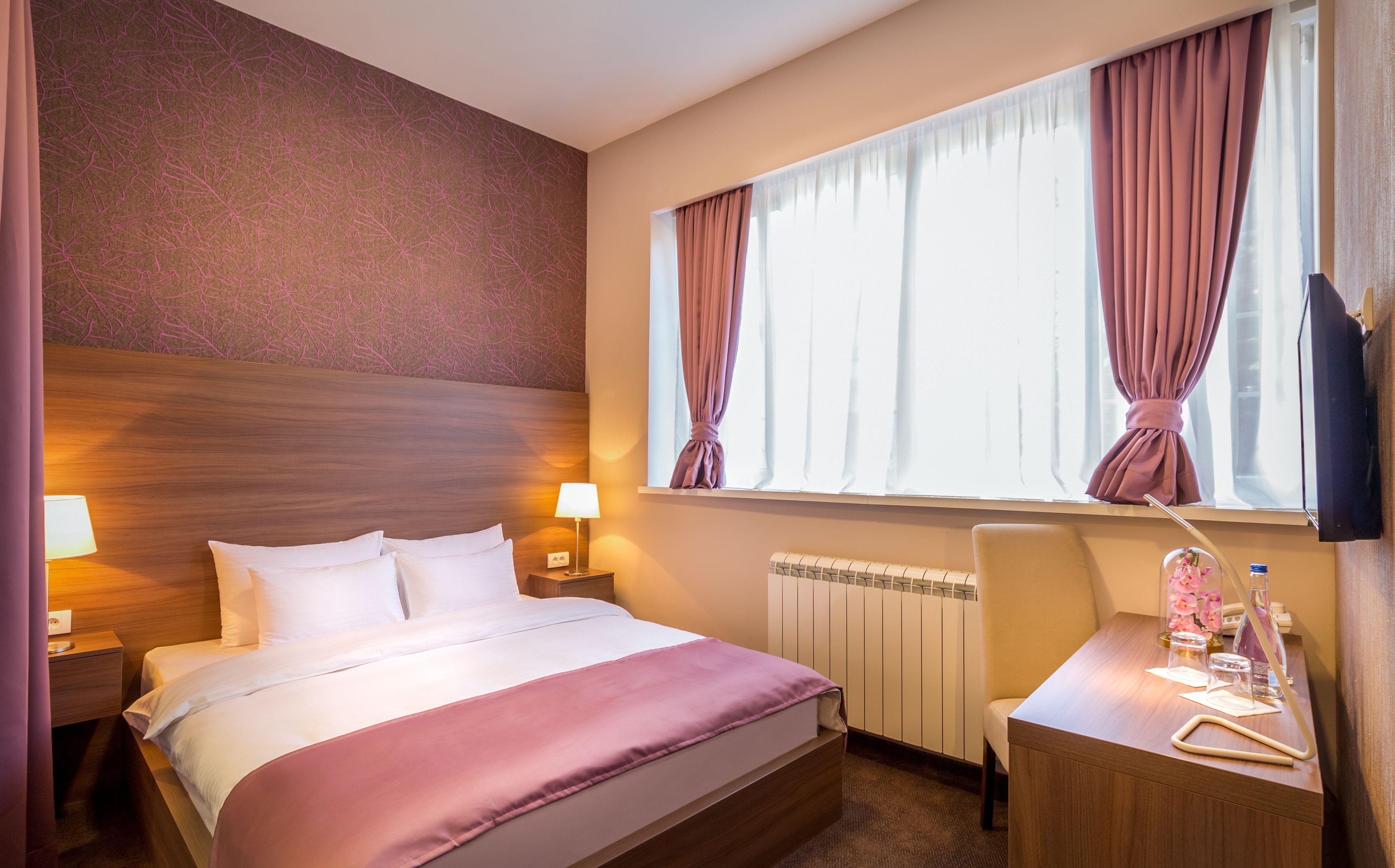Habitación de hotel con cama, lámparas de noche, un escritorio con silla, radiador y ventana con cortinas.