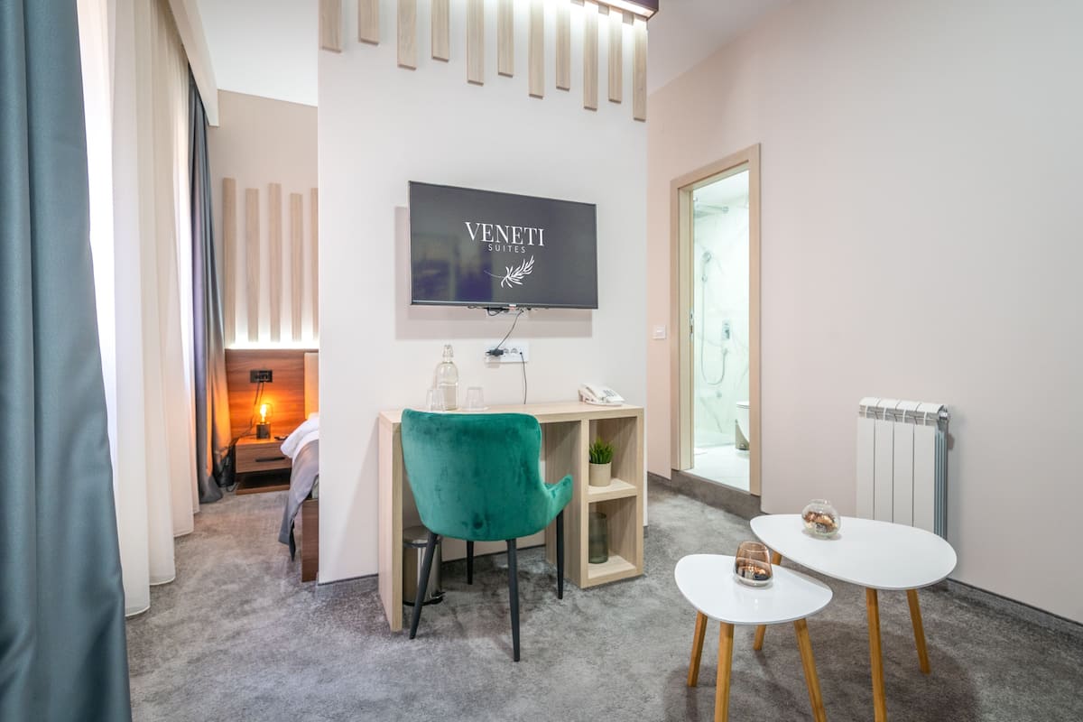 Ein modernes Hotelzimmer mit flachbildfernseher an der Wand, einem petrolfarbenen Sessel, einem kleinen weißen Tisch und einem Holzbüro mit Lampe.