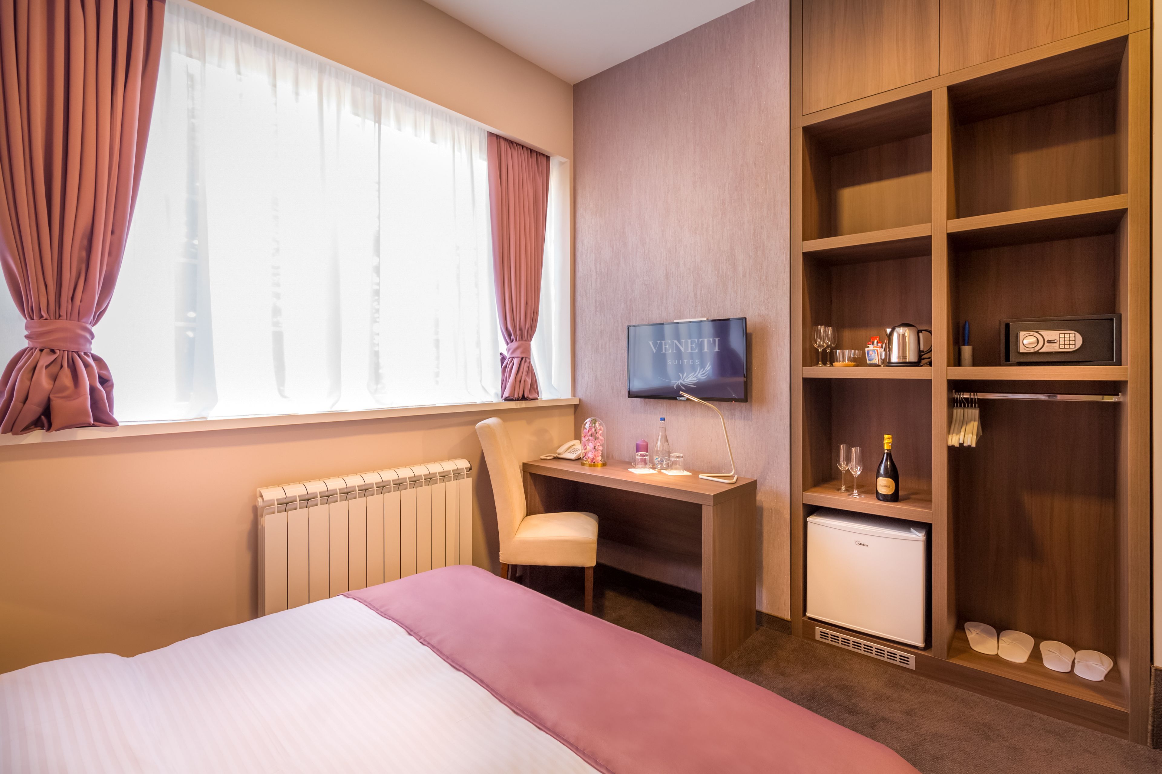 A hotel room with a bed, a desk with a computer, a TV, a mini-fridge, and a wardrobe.
