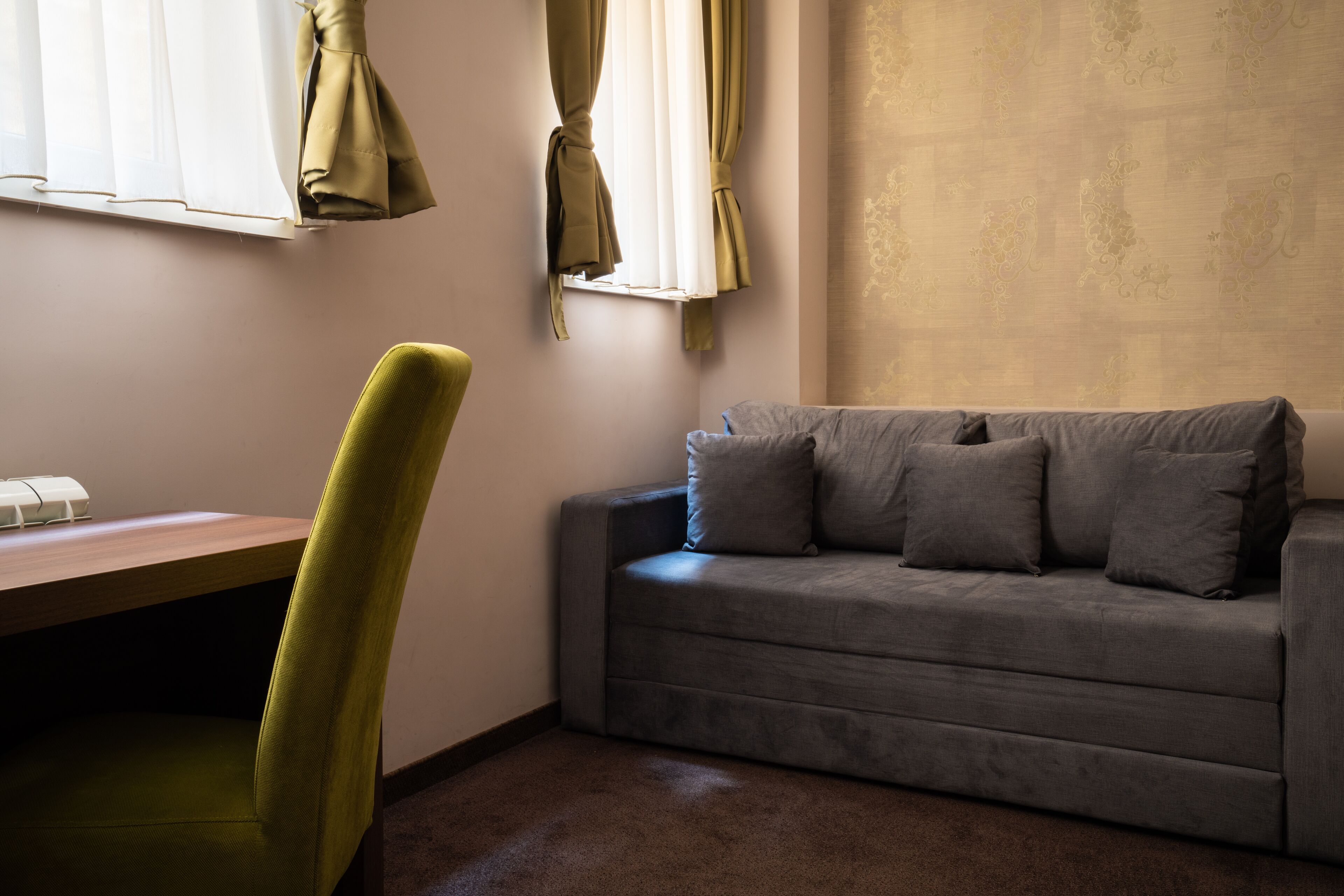 A room with a grey sofa, a green chair, a wooden desk, and a window with curtains.