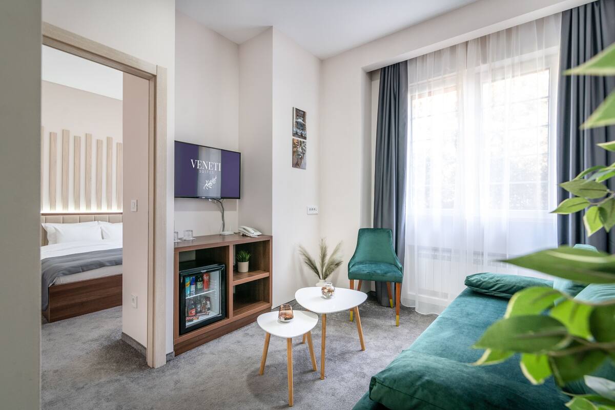 Ein Hotelzimmer mit Bett, Fernseher, kleinem Tisch, grünem Sessel und großem Fenster mit Vorhängen.