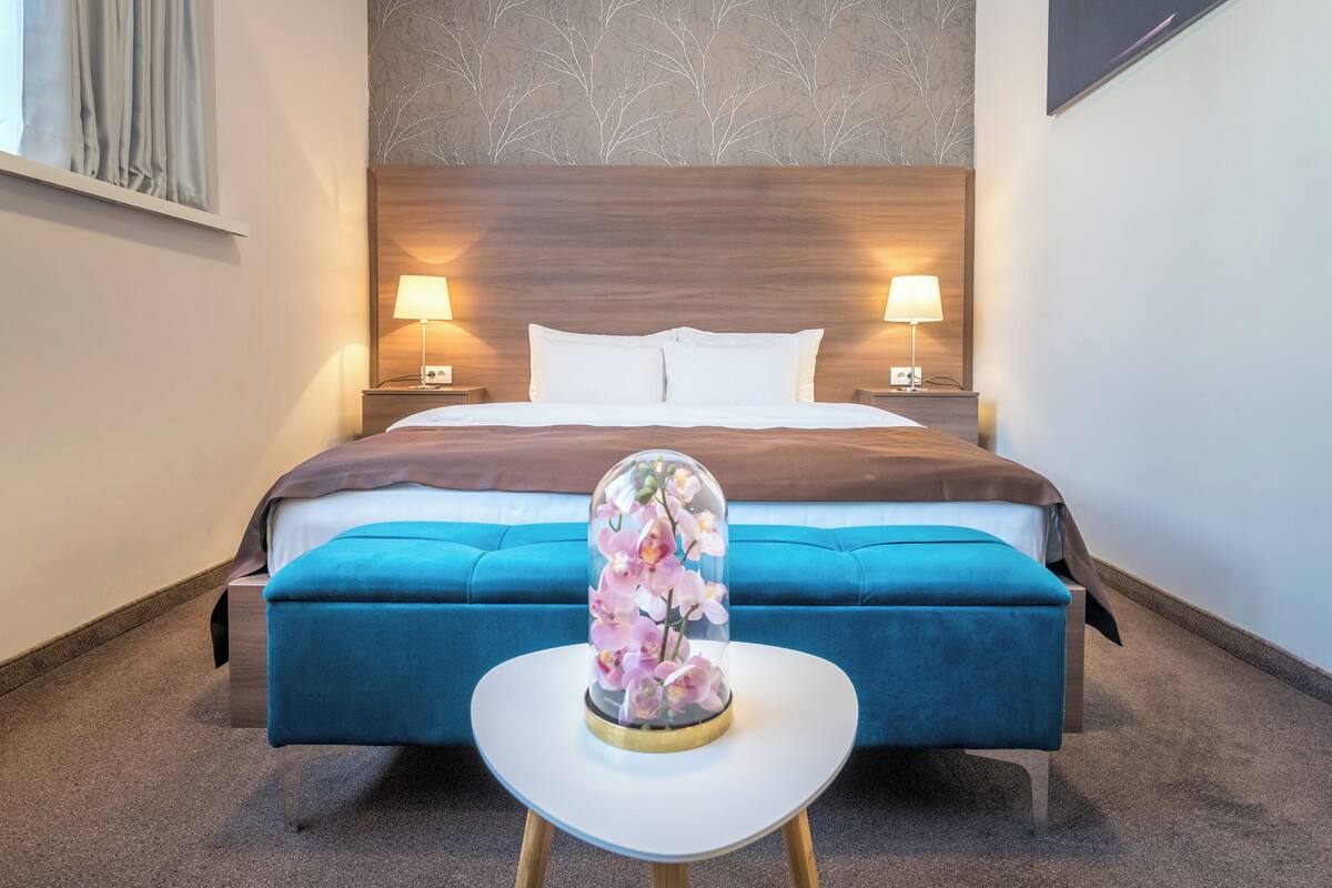 Ein Hotelzimmer mit einem großen Bett, zwei Nachttischlampen, einer blauen Sitzbank und einer Glaskuppel mit Blumen.